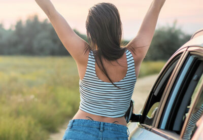 Een persoon in een gestreept shirt en jeans strekt zich uit naast een auto op een landweggetje en geniet van het uitzicht op de zonsondergang, alsof hij een vergeten vakantie herbeleeft.